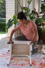 Hispanic woman kneeling on porch drop cloth sanding stool with sanding block next to paint can