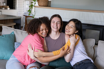 Naklejka premium Diverse female friends hugging on sofa in home living room, laughing with fireplace backdrop