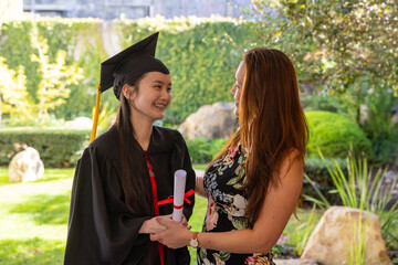 Asian mother and daughter celebrating graduation in courtyard holding diploma scroll and cap