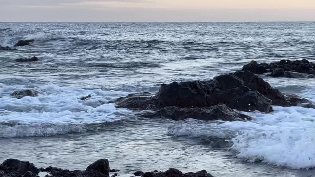 Calm ocean waves washing a rocky shore at dawn, soft pastel blue light before sunrise, minimal coastal scenery conveying freedom, travel lifestyle, nature balance, environment and relaxing seaside mor