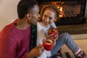 Naklejka premium Diverse couple sitting on rug before hearth fireplace with red mugs and draped fleece blanket