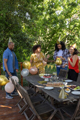 Diverse friends gathering around table on backyard deck with cake, party hats, metallic balloons