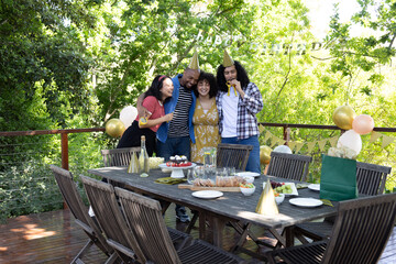 Diverse friends wearing cone party hats hugging and smiling on deck around birthday cake