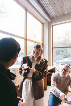 Young female design expert showing ideas on smart phone to coworkers in office