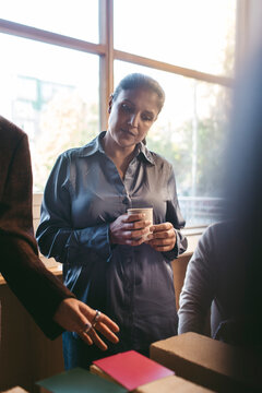 Mature businesswoman holding mug listening to colleague explaining architectural plan in office