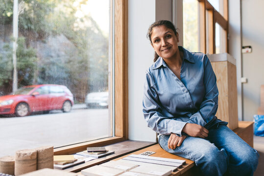 Portrait of smiling mature female architect sitting on window sill near wood samples in office
