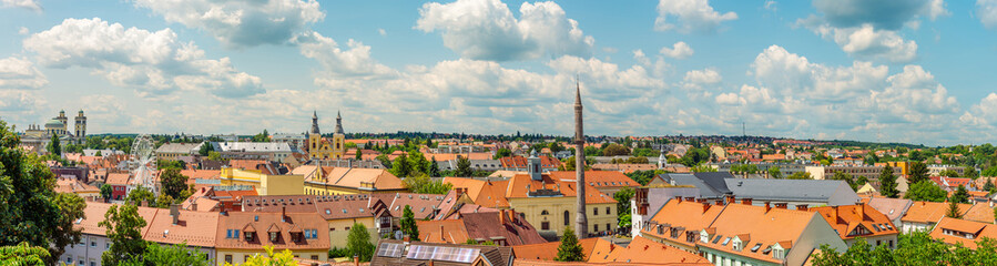 Obraz premium Expansive panoramic view of Eger, Hungary, showcasing a beautiful tapestry of red and orange terracotta-tiled roofs under a bright, cloud-filled summer sky. 
