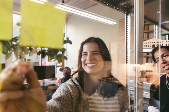 Smiling female design professional writing pointers on glass door during meeting in new office