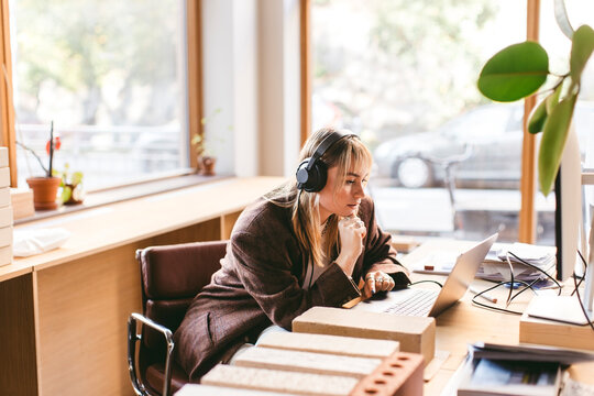 Concentrated young design professional wearing wired headphones using laptop while working in office