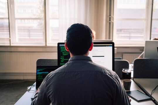Rear view of young male programmer working on computers while sitting at desk in tech office