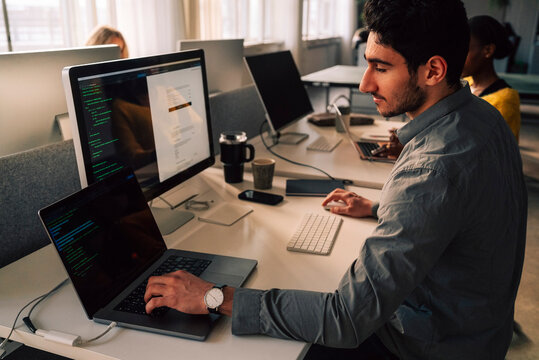 Young male programmer typing on laptop while sitting at desk in tech office