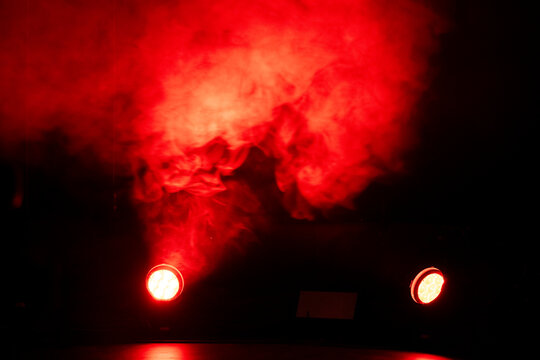 Bright red lights pierce through swirling smoke in a dimly lit space, creating an intense atmosphere during a live performance or event at night.