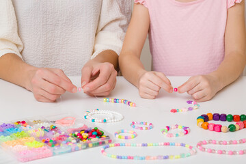 Mother and little child girl hands creating beaded jewellery from different colorful plastic parts on white table at home room. Closeup. Front view.