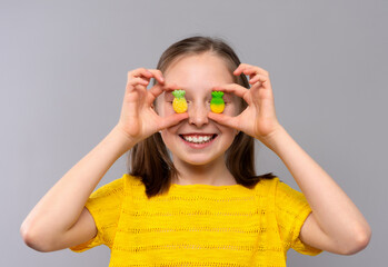 A cheerful young girl with light skin and brown hair in pigtails is posing against a light gray studio background. She is wearing a bright yellow knitted shirt and smiling broadly. The child playfully
