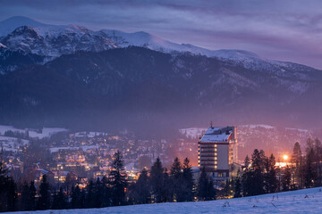 Night cityscape of Zakopane with the silhouette of Tatra Mountains. Illuminated mountain resort town in Poland under a dark evening sky.