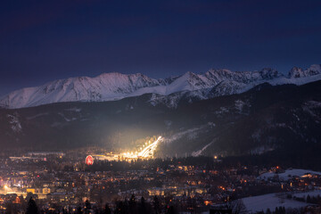 Night cityscape of Zakopane with the silhouette of Tatra Mountains. Illuminated mountain resort town in Poland under a dark evening sky.