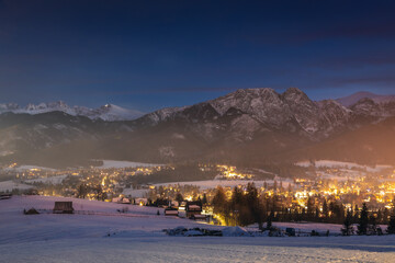 Night cityscape of Zakopane with the silhouette of Tatra Mountains. Illuminated mountain resort town in Poland under a dark evening sky.