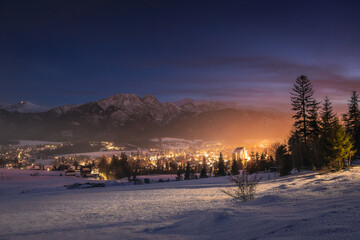 Night cityscape of Zakopane with the silhouette of Tatra Mountains. Illuminated mountain resort town in Poland under a dark evening sky.
