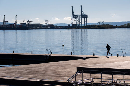 Harbor cranes rise over waterfront boardwalk as silhouette runner keeps run under open sky with urban industry creating calm scene