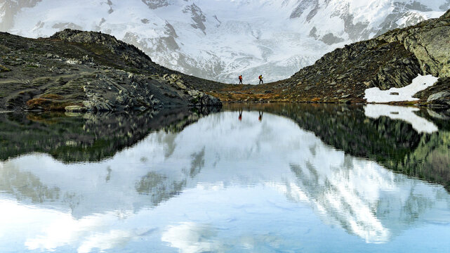View of two hikers journeying across a rocky ridge, mirrored in the serene lake below, with snow-capped mountains in the distance, Zermatt, Valais, Switzerland.