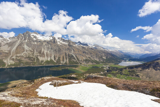 View of snow-capped mountains reflected in the lake's dark waters under a bright blue sky dotted with fluffy white clouds, Maloja, Grisons, Switzerland.
