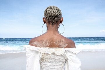 Back view of a woman with short textured hair and hoop earrings standing on a tropical beach, looking at the ocean. Minimalist summer scene symbolizing freedom, calm, travel, self-reflection, and conn