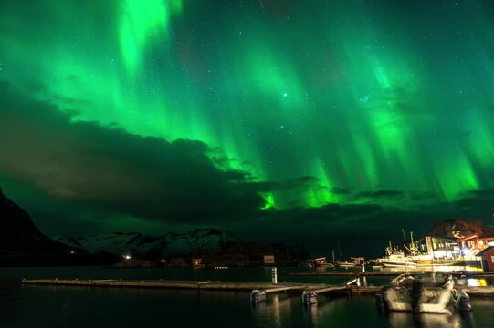 Massive Northern lights (aurora borealis) KP3 appearing green over the sky at a dock with boats in Senja island, Norway
