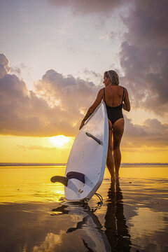Silhouette of a young woman carrying a surfboard and walking into the ocean at sunset. Scenic seascape with warm evening light symbolizing freedom, adventure, surf lifestyle, travel, and connection wi