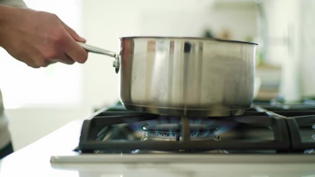 Clean, well-groomed hands in a neutral shirt position a sleek stainless steel saucepan onto a pristine induction hob in a minimalist kitchen, with warm light and steam, a macro close-up for cinematic
