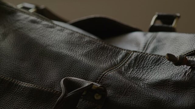 Close-up footage of hands pulling metal zipper on dark leather bag. Shallow depth of field emphasizes texture of natural leather, stitching, and the zipper mechanism. Travel preparation and packing