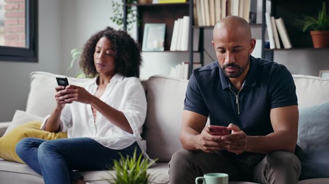 Video of angry couple sitting on couch together and looking to opposite sides while using smartphone at home.