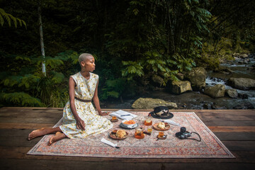 A dark-skinned woman sits on a rug, enjoying a picnic in the tropical jungle near a mountain stream. She wears a stylish dress, blending fashion with natural surroundings. 