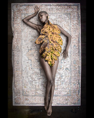 A dark-skinned woman lies on a rug, covered by a single large tropical leaf, photographed from a top-down perspective. The scene combines natural aesthetics with a fashion-inspired concept, highlighti