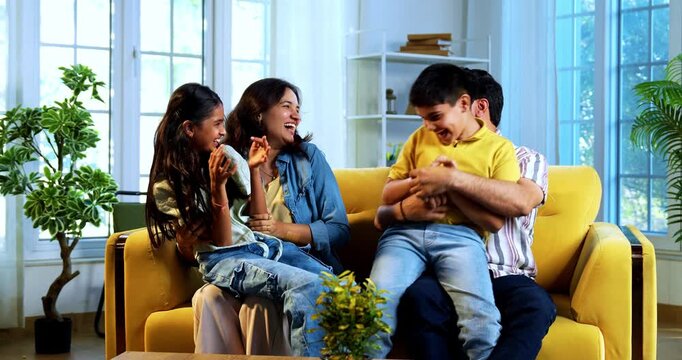 Indian young parents tickling kids at home while sitting on sofa in playful living room, happy father and mother laughing with son and daughter indoors, joyful family lifestyle moment