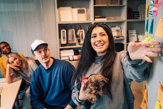 Smiling young businesswoman carrying dog while explaining pointers through adhesive note to team in tech office