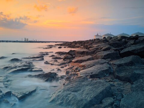 A beautiful, warm-toned photograph of a rocky shore as the sun sets or rises over the ocean. The sky is filled with soft orange, pink, and blue hues, reflecting gently on the water