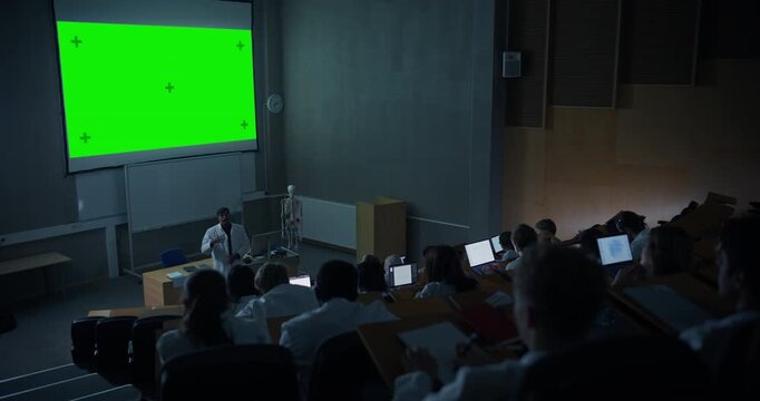 Male Professor in Lab Coat Lectures Medical Students in a University Auditorium, Uses Large Green Screen Chroma Key Projector With Tracking Markers. Mock-up Content Concept. Static Shot