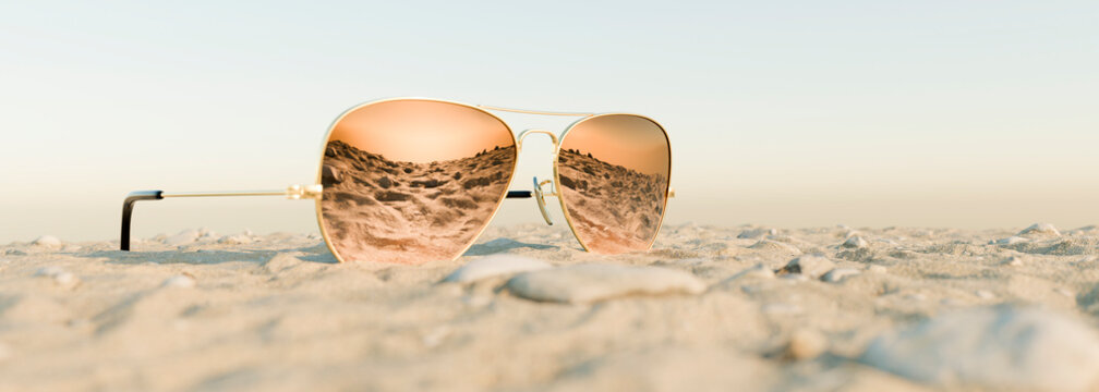 Pair of modern sunglasses on a sandy beach on a clear blue sky day with a sunset beach reflected in the glasses 3d render