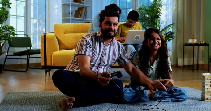 Indian father daughter playing video game and mother son sitting on sofa behind as family enjoys fun time together indoors in modern lavish home living room lifestyle scene