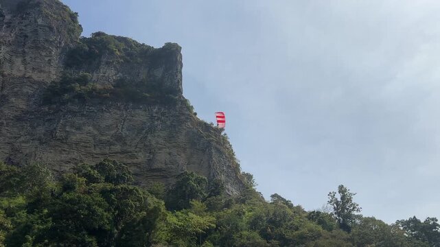 Parachutist descending with open canopy beside tall limestone cliff and forested hillside near Ao Nang Beach, aerial action scene under clear daylight sky in Krabi Thailand