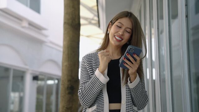 Excited young woman checking smartphone and celebrating good news outside modern building. Success and achievement concept.
