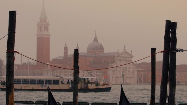 Vaporetto water bus transports passengers across the Venetian lagoon on a hazy day. San Giorgio Maggiore church silhouette visible in background with wooden mooring piles in foreground.