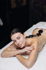 Female model receiving a spa treatment with smooth black stones placed along her back, lying on a white massage table in a serene spa environment
