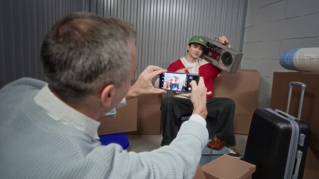Father photographing his son with smartphone in self storage room, young man sitting on box and posing with vintage ghetto blaster on his shoulder