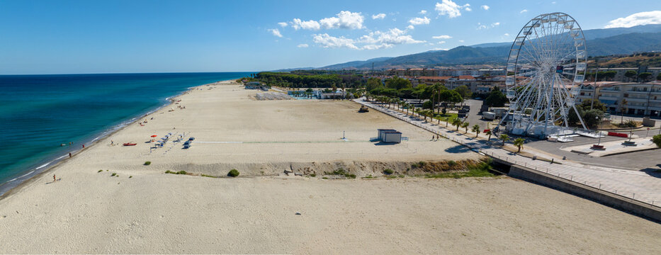 Aerial view of a vast beach and coastal town, featuring a large white ferris wheel on the right, a long stretch of golden sand, and the deep blue sea under a clear sky. It is the seafront of Soverato.