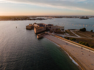 Aerial view of the Citadelle de Port-Louis in Brittany, France, glowing in the warm golden hour light of sunset, highlighting its fortifications, bastions, and coastal position along the harbor.

