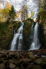 Portrait view of the Cascade du Heidenbad in the Vosges Mountains, showing cascading water flowing over dark rock faces, surrounded by forest vegetation and a calm, natural atmosphere.

