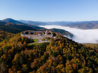 Wide aerial view of Ch&acirc;teau du Hohlandsbourg atop a hill, overlooking a valley filled with low white clouds beneath a clear blue sky, highlighting its dramatic Alsace landscape setting.

