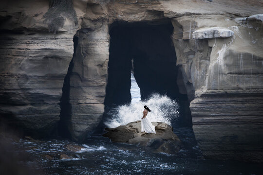 Distant side view of a mid adult Woman in a long dress standing on coastal rocks with waves crashing around her, California, USA