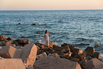 Wide shot of a young woman standing on a scenic rocky beach by the blue ocean water